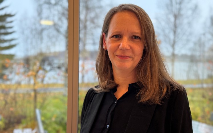 Caroline Pfriender står vid ett fönster, höstlandskap i bakgrunden.