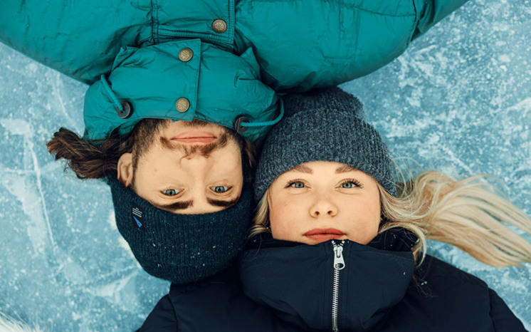 Närbild på två vinterklädda unga vuxnas ansikten som ligger omlott på isen och ser in i kameran.