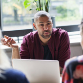 En man sitter vid ett bord med en bärbar dator framför sig och pratar med kollegor