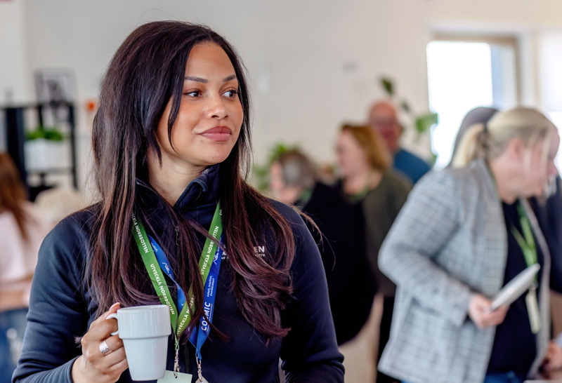 Isa Ponturo står med nyckelband runt halsen och en kaffekopp i handen, workshopdeltagare i bakgrunden.