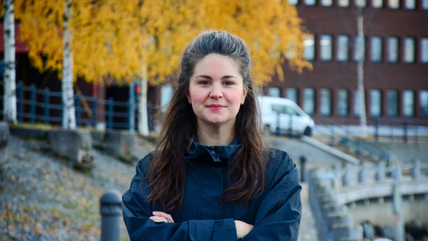 Kvinna med långt mörkt hår står med armarna i kors utanför en röd tegelbyggnad.