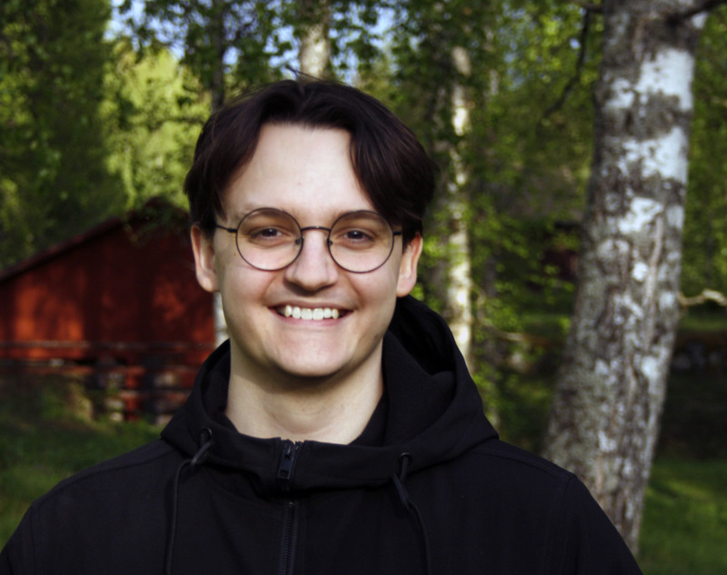 Maximilian Östlund står ute i naturen med en röd byggnad bakom sig.