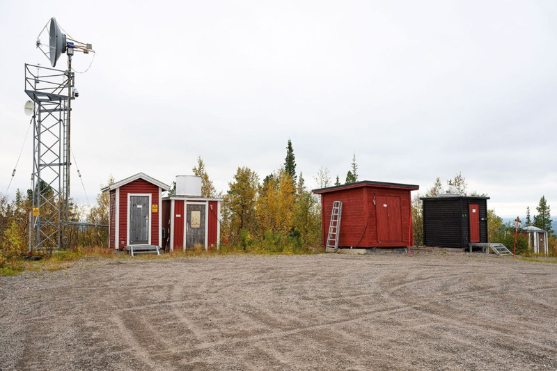 En grusplan med små faluröda bodar i trä. En ställning med en antenn syns till höger.