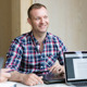 Leende man i rutig skjorta sitter framför dator.