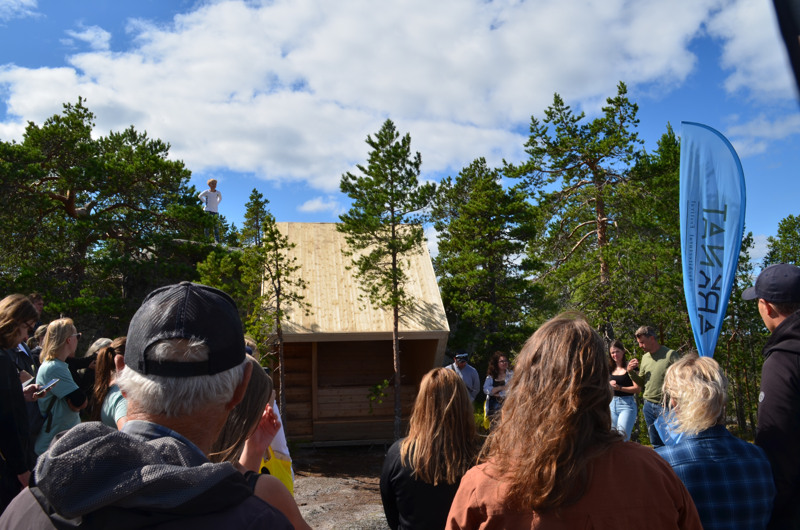 Besökare vid ett av vindskydden i samband med invigningen. Människor, skog, ett vindskydd och beachflagga för Arknat.