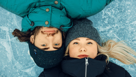 Närbild på två vinterklädda unga vuxnas ansikten som ligger omlott på isen och ser in i kameran.
