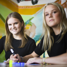 Två unga personer med långt hår sitter och ritar eller skriver framför en färgglad bakgrund.