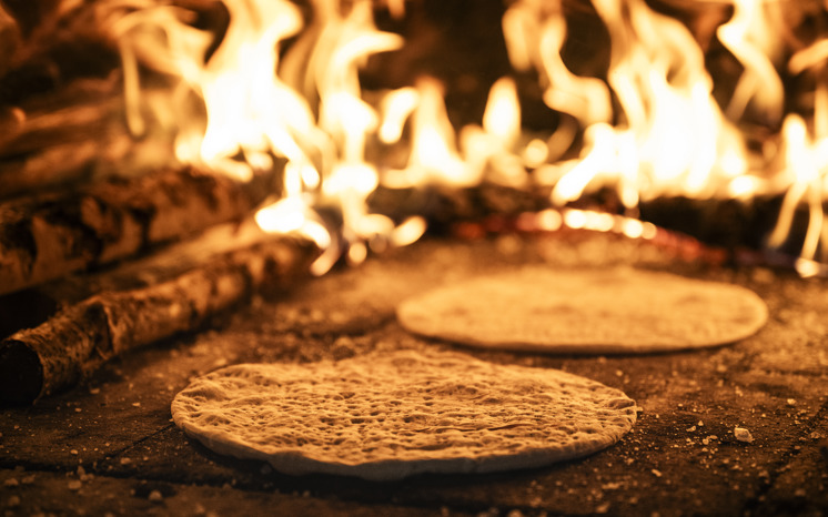 Två rieskakakor på gräddning i vedugn med eldsflammor i bakgrunden. 