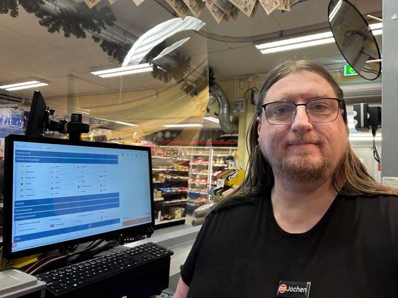 En man med långt hår och glasögon står framför en självscanningskassa i en dagligvarubutik.
