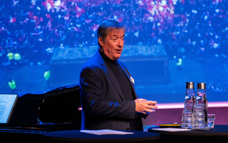 Neil Peterson på scen under Kulturkraft, visar bild av en folksamling i Liverpool på skärm bakom sig.